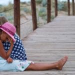 girl in blue and white polka dot shirt and pink hat sitting on wooden bridge