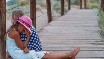 girl in blue and white polka dot shirt and pink hat sitting on wooden bridge